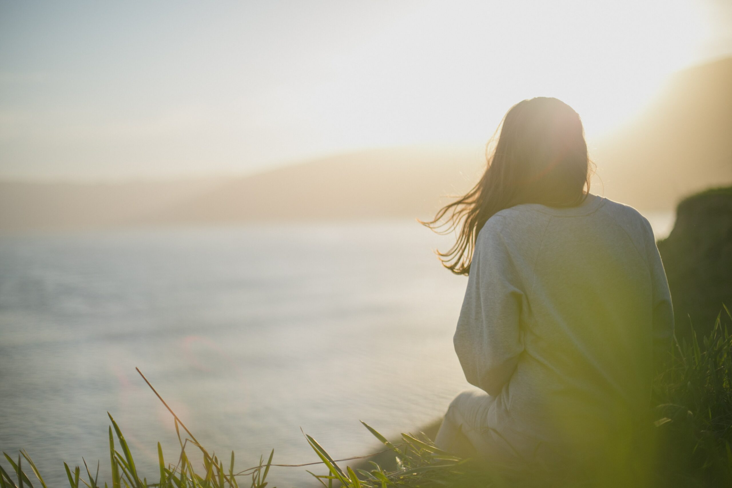 The back of a woman, who is sitting on a grassy hill overlooking a body of water